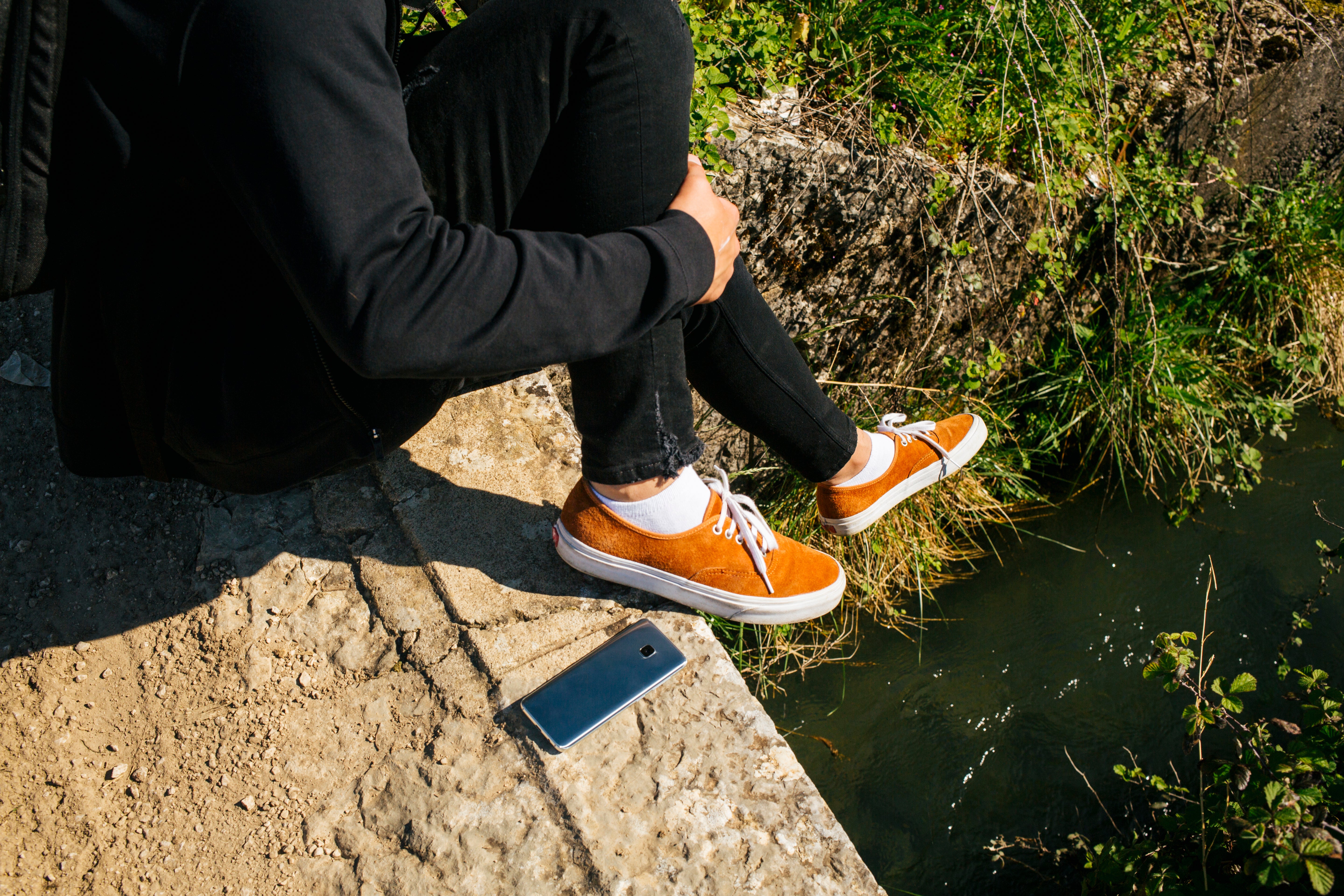 Person sitzt an einem Bach. Der Fokus liegt auf den Füßen. Person trägt Sneaker Socken.