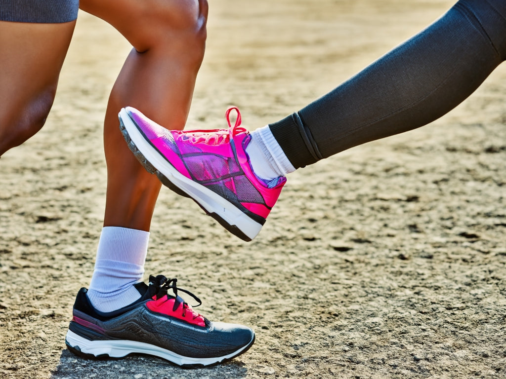 Zwei Sportler mit Sportsocken und Laufschuhen beim Training auf einer sandigen Laufstrecke. Weiße, atmungsaktive Sportsocken kombiniert mit schwarz-roten und pinken Laufschuhen. Perfekte Feuchtigkeitsregulierung und Blasenschutz für Läufer.