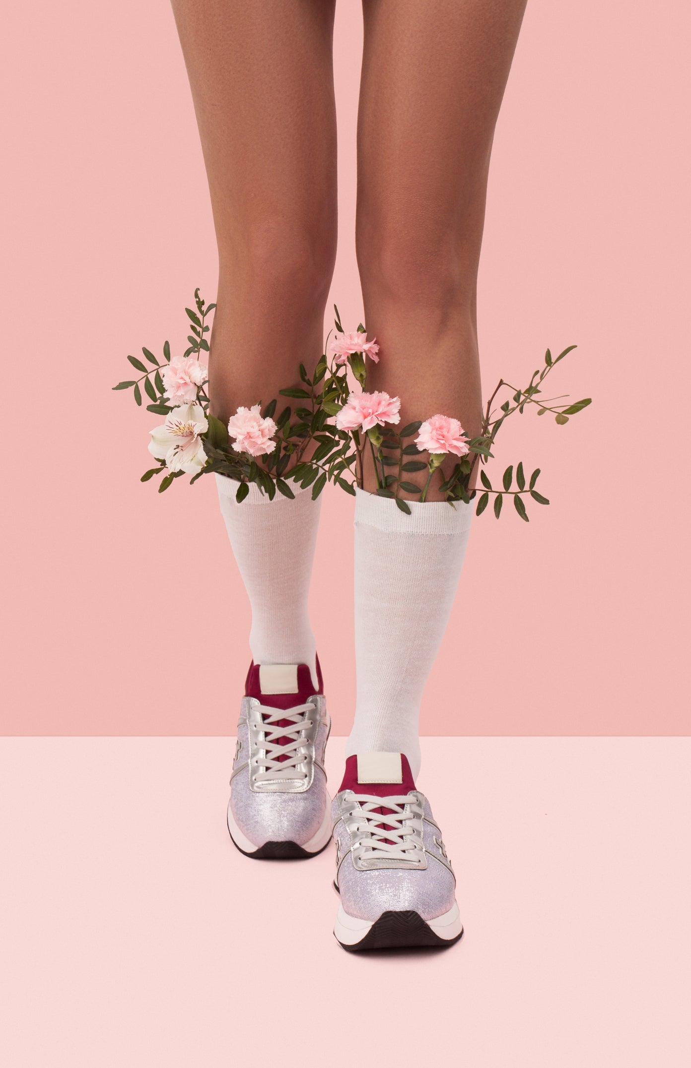 Person trägt weiße Kniesocken und glänzende Sneakers, dekoriert mit Blumen um die Knie, auf einem pastellfarbenen Hintergrund. Der Fokus liegt auf den Beinen und dem floralen Arrangement in den Socken.