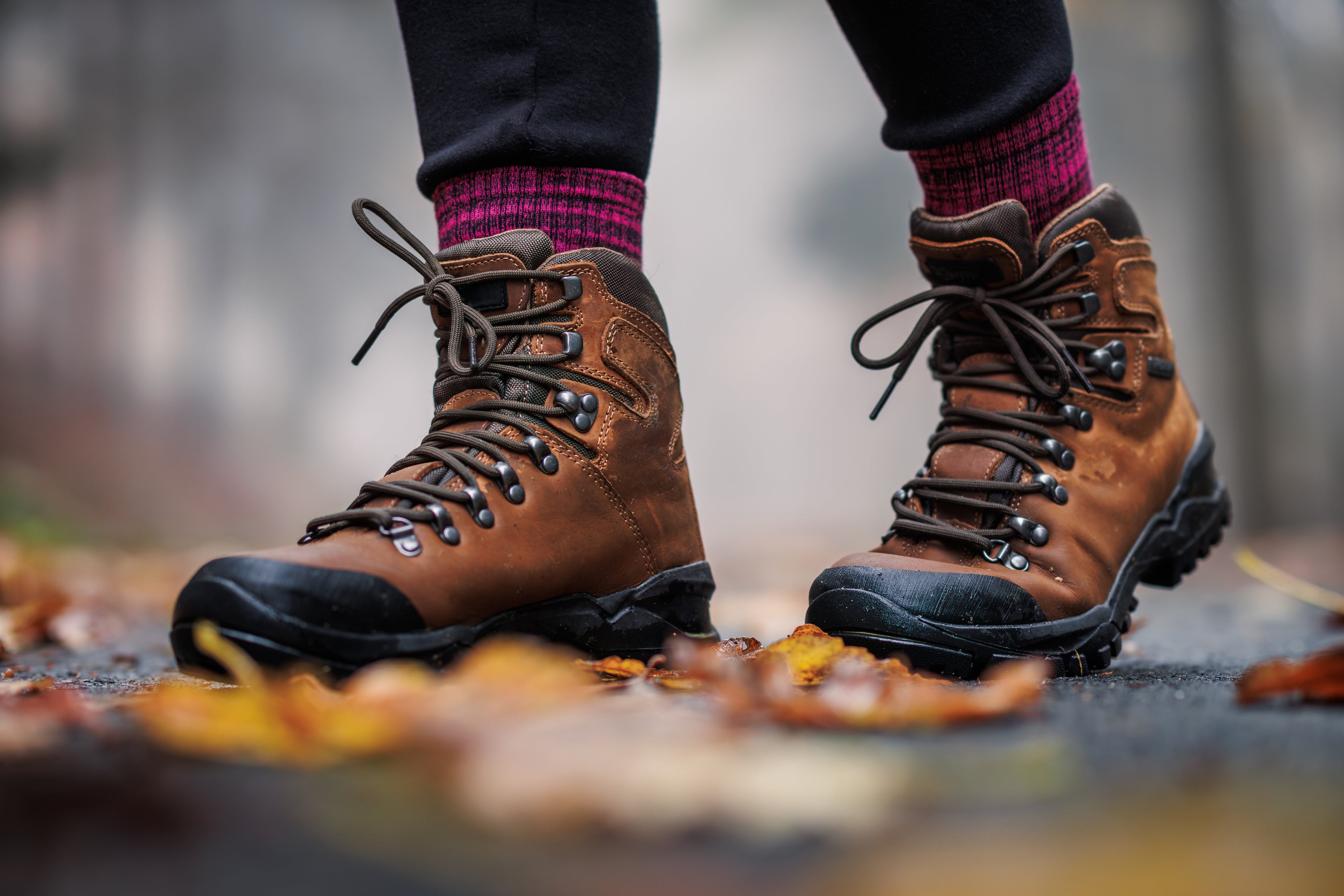 Eine Person trägt braune Wanderschuhe und warme, violett gemusterte Wollsocken, steht auf einem herbstlich bedeckten Weg mit nassen Blättern. Der Fokus liegt auf den robusten Schuhen und den wärmenden Socken, ideal für Outdoor-Abenteuer